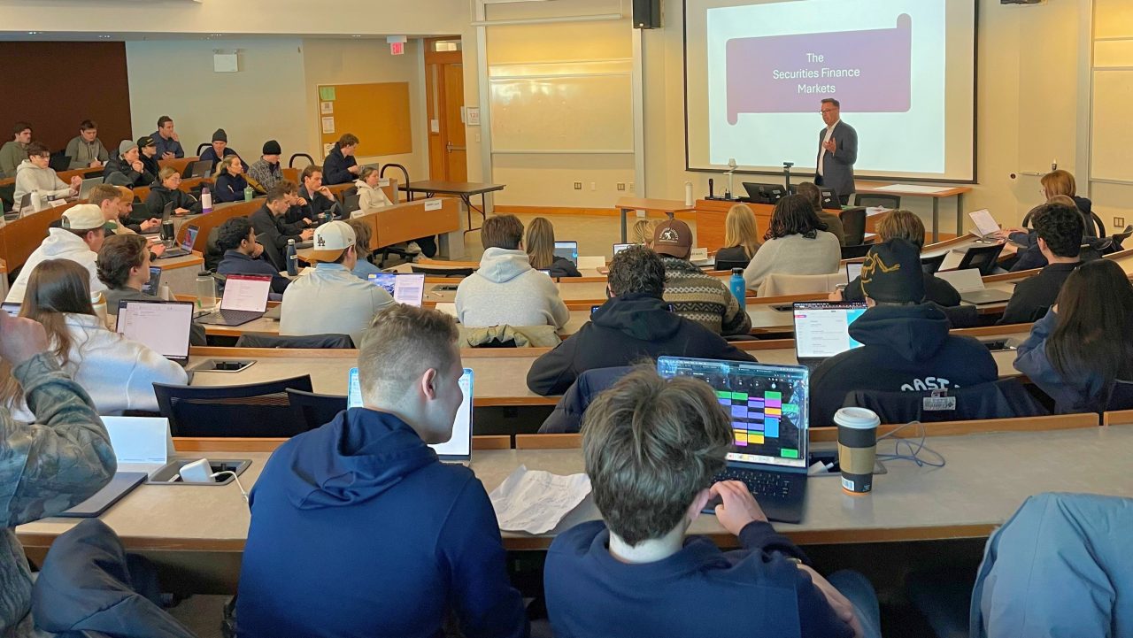 Students sitting in a lecture theatre working on laptops and focused on a person presenting and a screen at the front of the classroom.