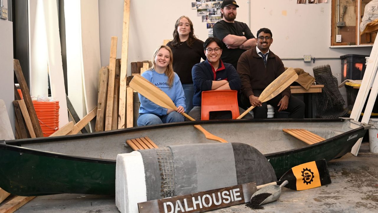 Five people sit and stand with two holding paddles, behind a cement canoe in a workshop.