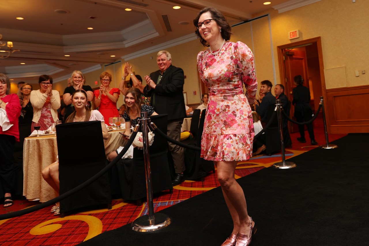 Kim Furlong walks along a black carpet while event attendees seated and standing cheer her on.