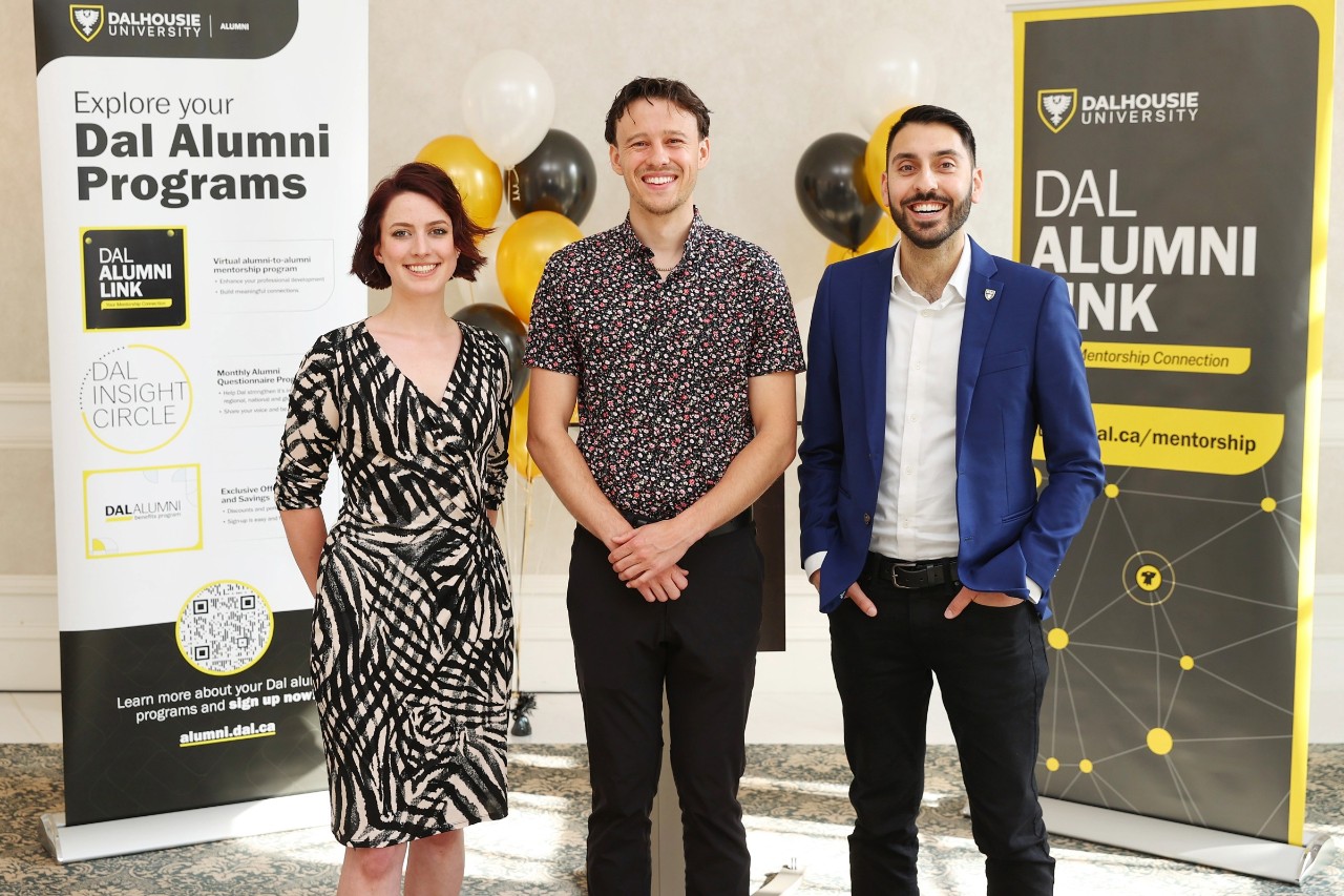 Three people dressed business casually smile at an event with banners behind them.