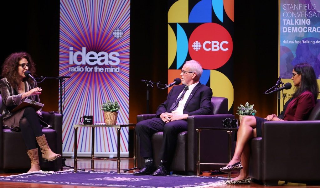 Two women and a man sitting on stage in soft seating with microphones in conversation at an event.