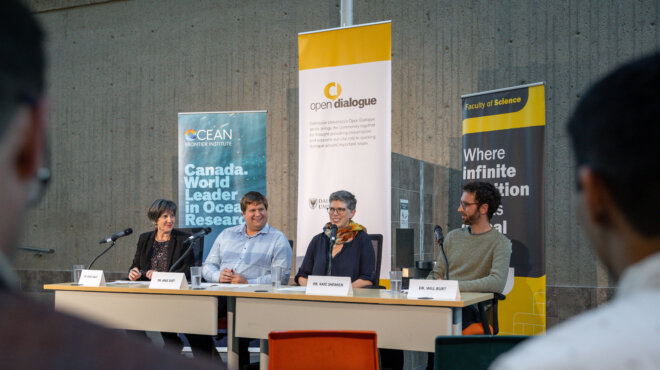 Four people sit at a table with microphones in front of event banners with attendees in the foreground.