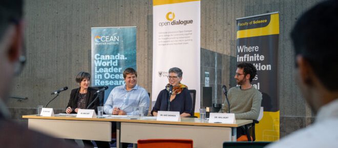 Four people sit at a table with microphones in front of event banners with attendees in the foreground.