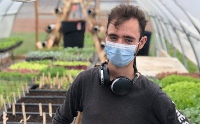 A man stands in a greenhouse with hands on hips, wearing a grey shirt, mask, and headphones around his neck.