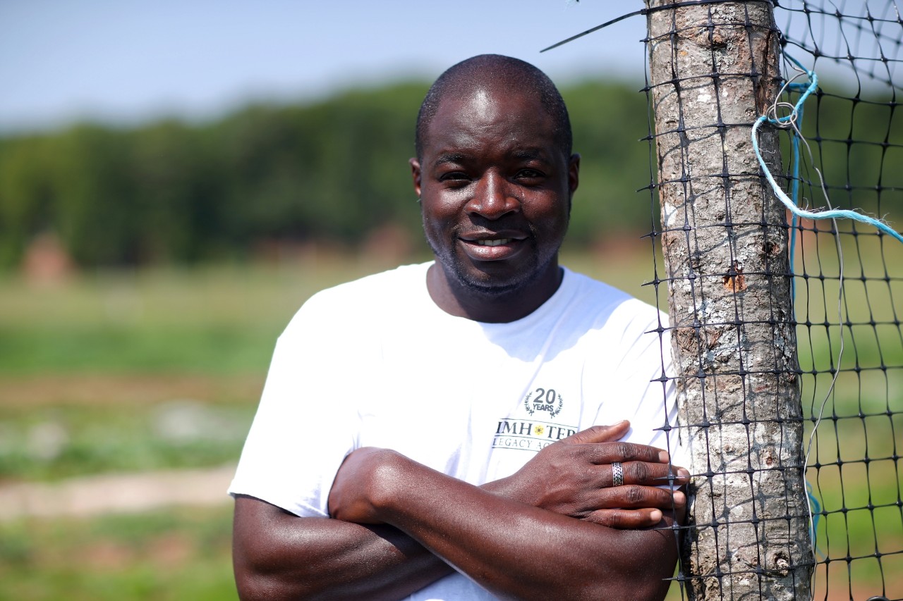 Foster leans on a fence post with his arms crossed on a farm in fine weather.