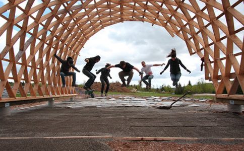 A group of 6 students jumping beneath an arched latticed structure  outside.