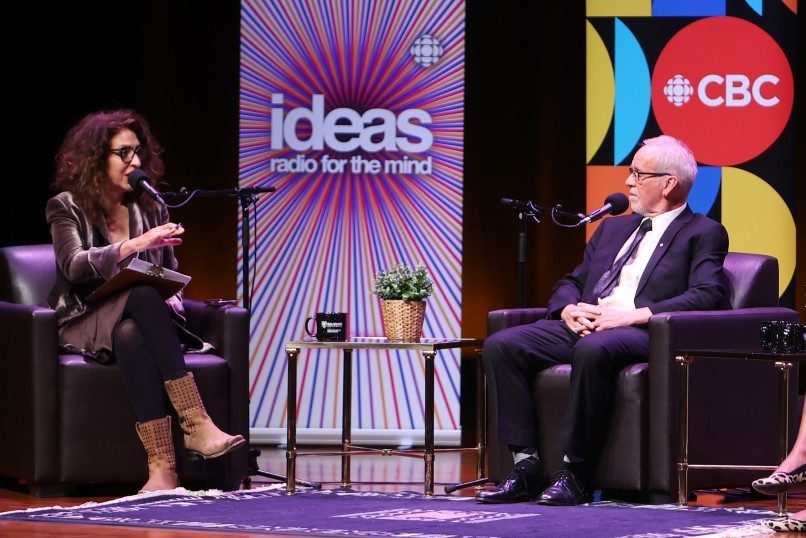 Two women and a man sitting on stage in soft seating with microphones in conversation at an event.