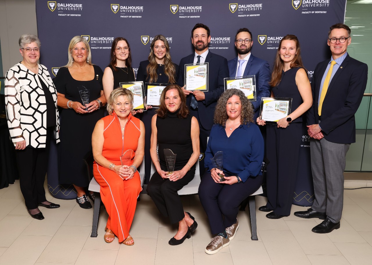 Award recipients stand holding framed certificates or crystal awards alongside several others standing and seated.