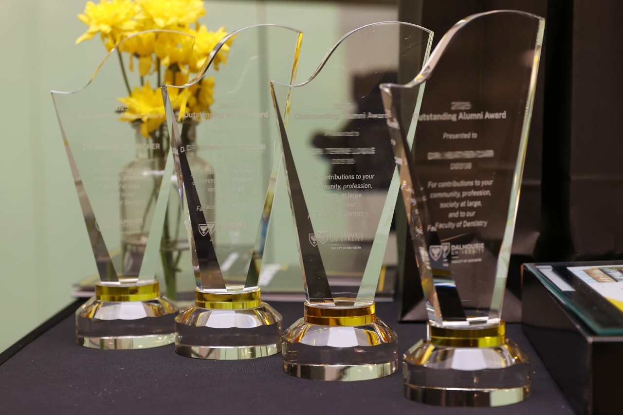 Four crystal awards etched with recipient information lined up on a table with a vase of yellow flowers.