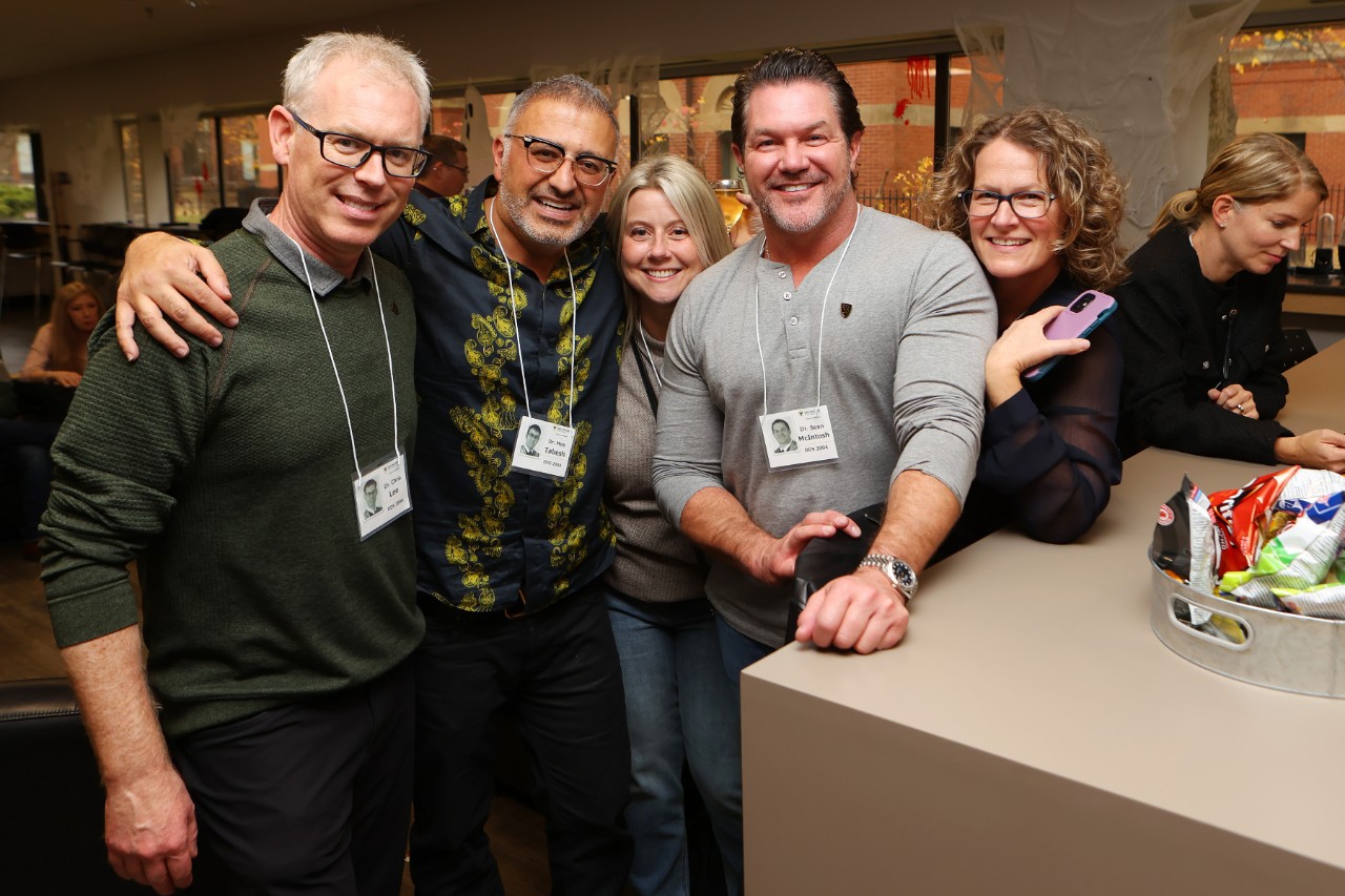 A group of 5 people stand close together at a casual get together smiling and wearing nametags on lanyards.