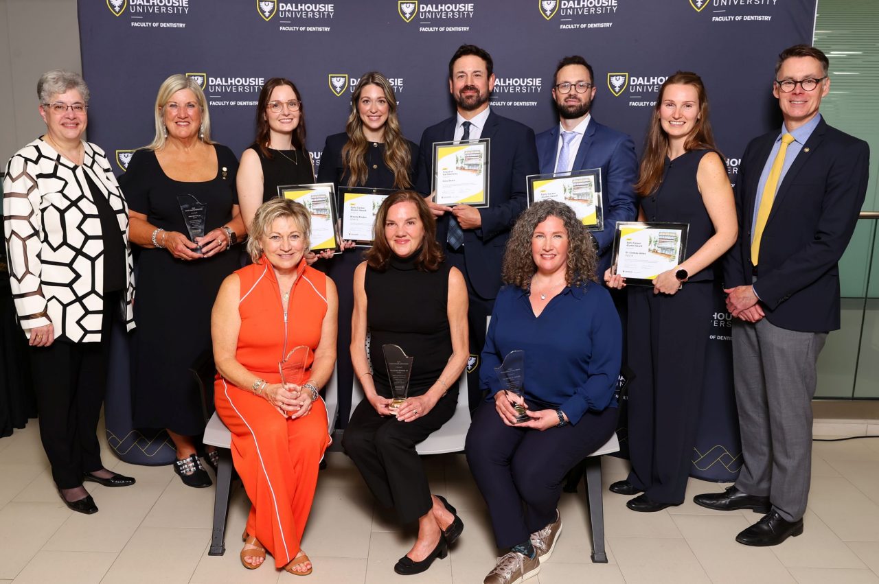 A group of people with a standing back row and three people seated holding crystal awards or framed certificates dressed for a semi-formal awards ceremony.