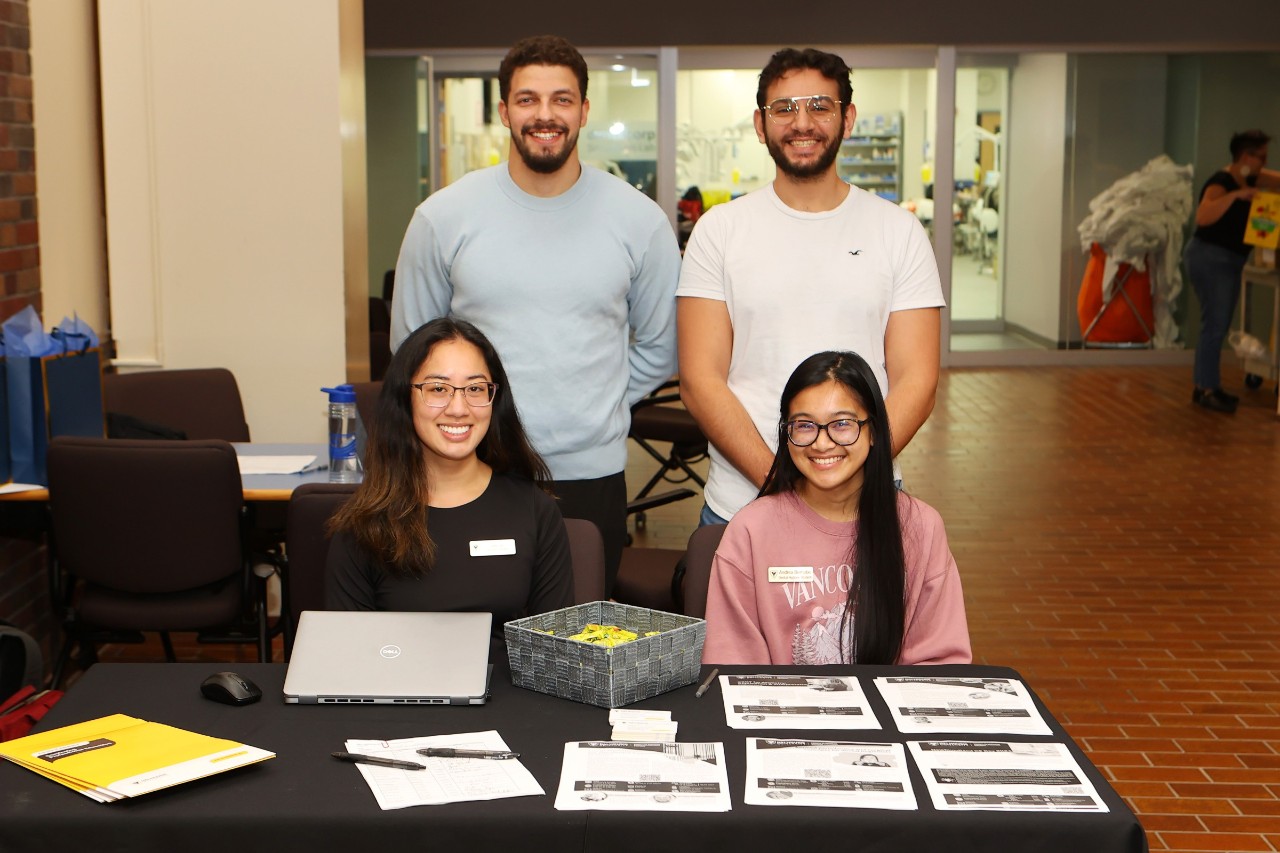 Two women are seated at a registration table with papers, pens and a laptop while two men stand smiling behind them.
