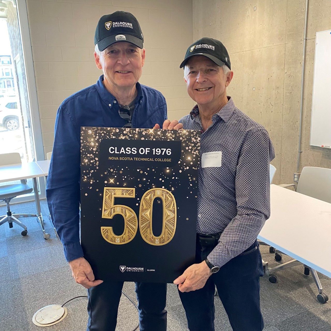 Two members of the engineering class of 1976 hold a sign with a gold "50" and wear Dalhousie ball caps.