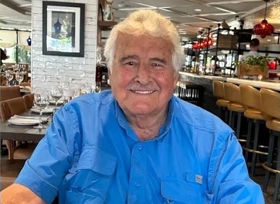 A man wearing a light blue shirt smiling in a restaurant set for semi-formal dining.