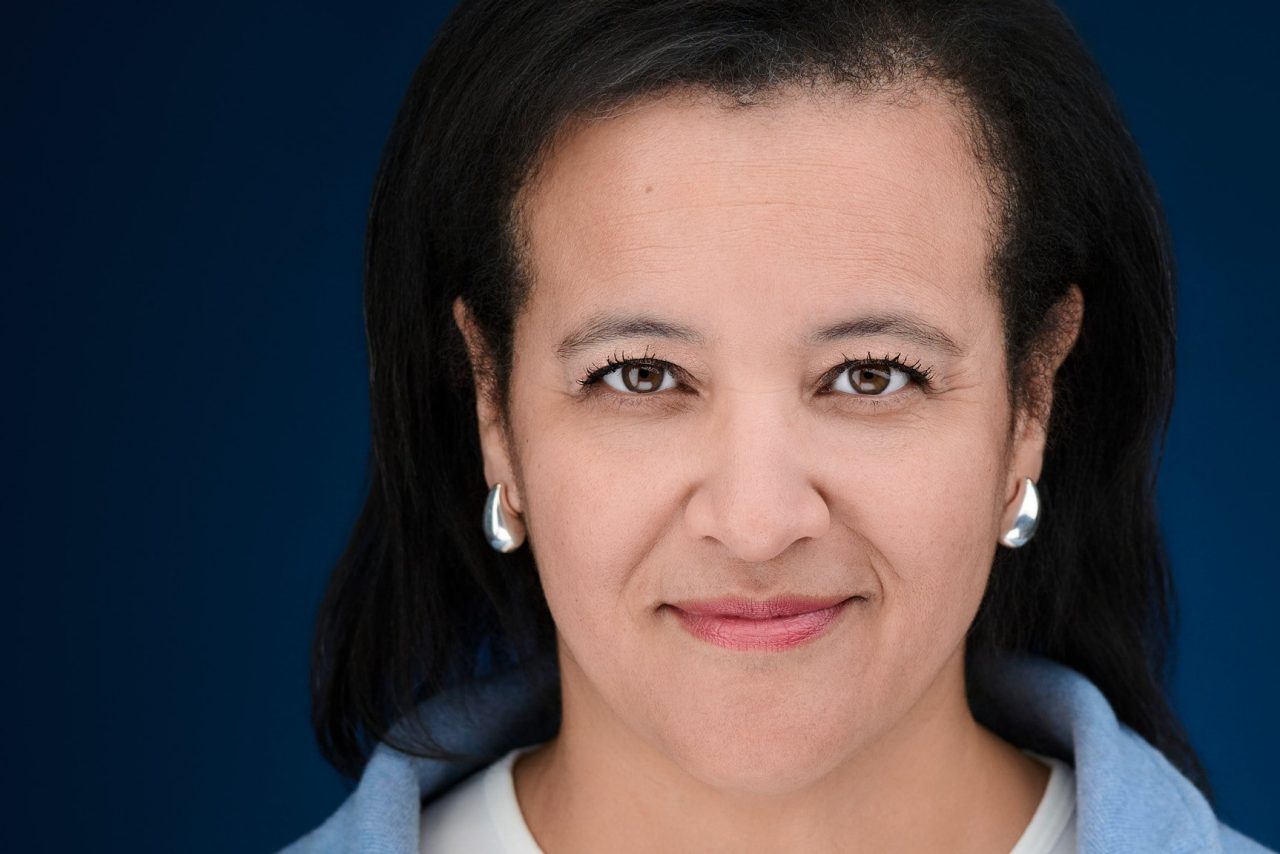 A woman wearing a white t-shirt and blue sweater looks at the camera