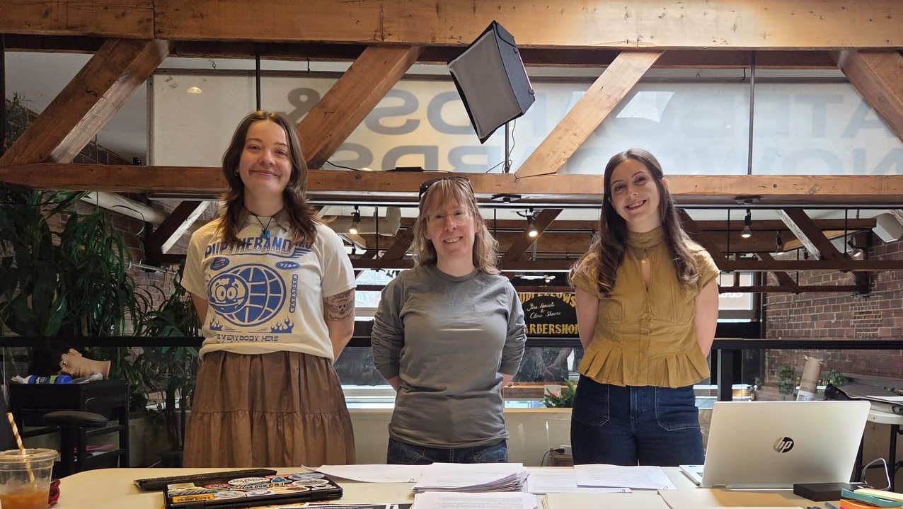 Three women stand behind a desk covered with papers and a laptop