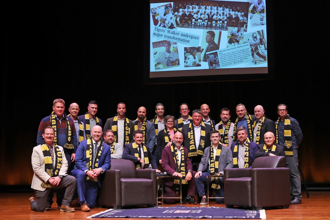 A group of former student-athletes wearing black and gold scarves pose on stage at an event.