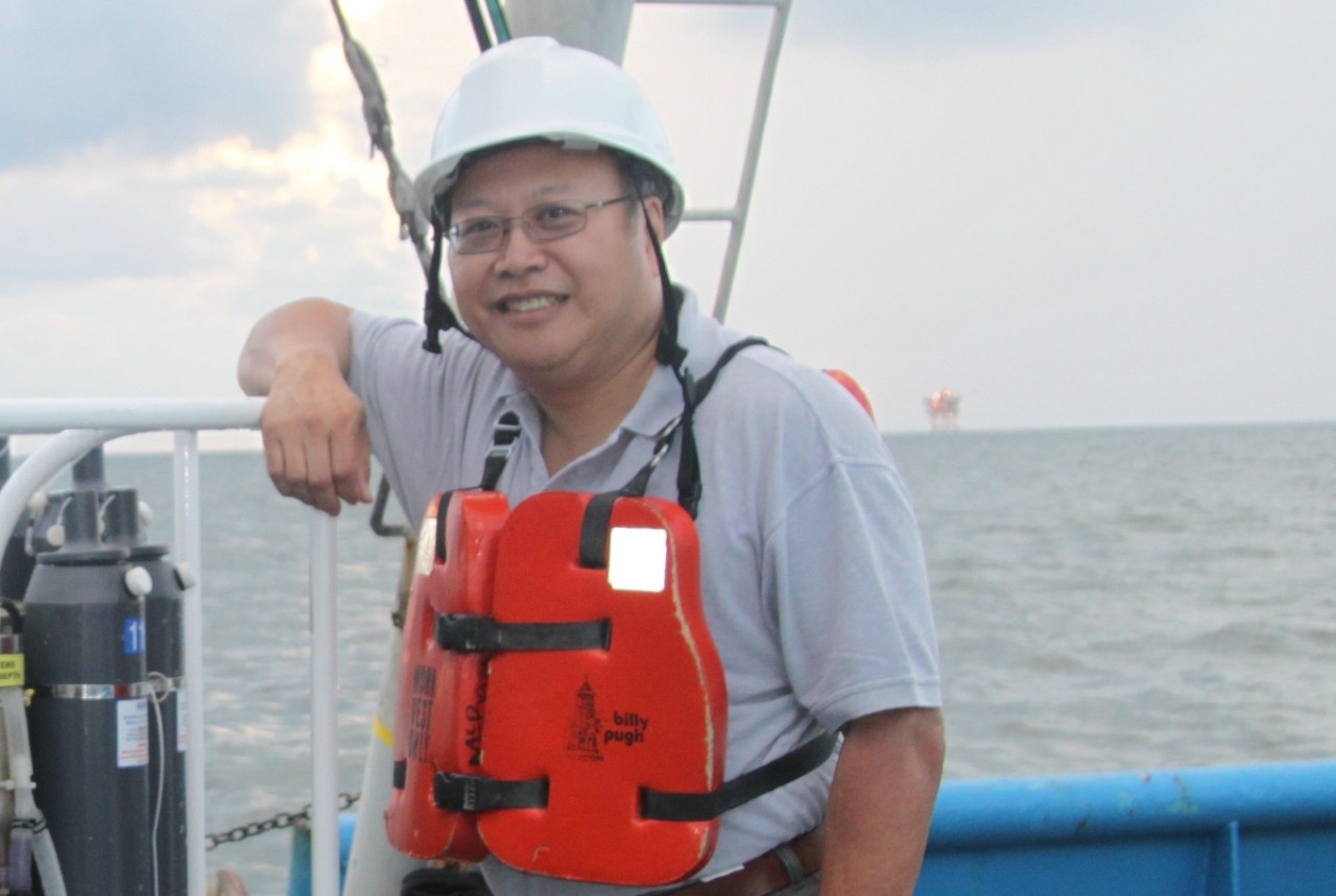 A man stands smiling aboard a ship at sea wearing a life preserver and a hard hat.
