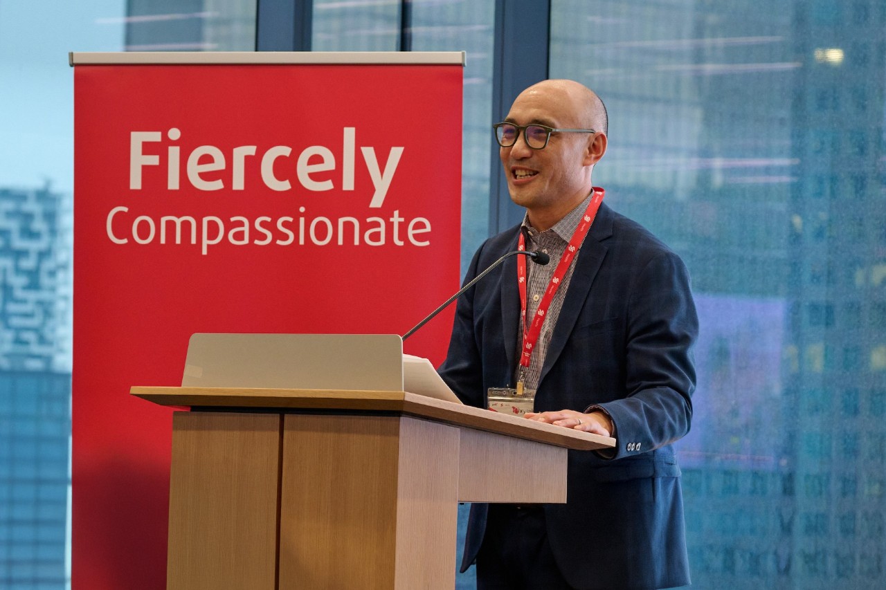Person speaking at a podium beside a red “Fiercely Compassionate” sign with large windows and a cityscape in the background.