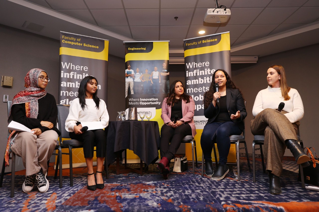 Five women of diverse backgrounds sitting in a row of chairs holding microphones and one is speaking at an event with pop-up banners in the background.