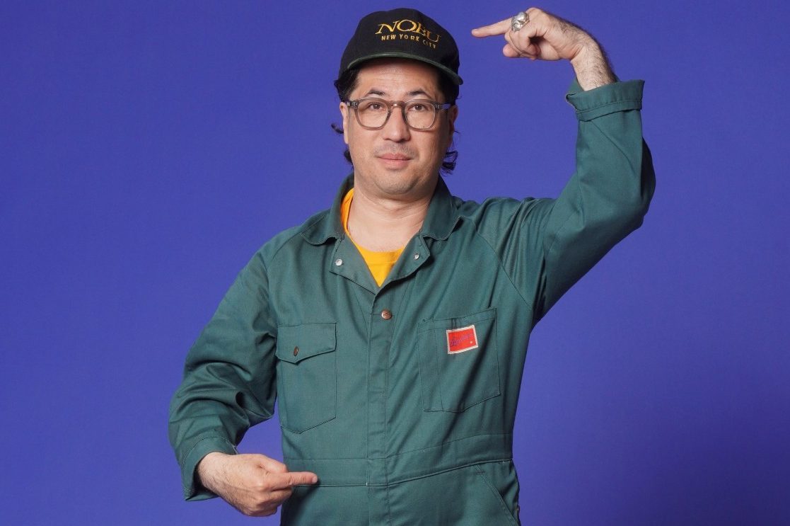 Nobu Adilman in a green jumpsuit and cap standing in front of a brown couch against a purple backdrop pointing to his head with one hand and midsection with the other.