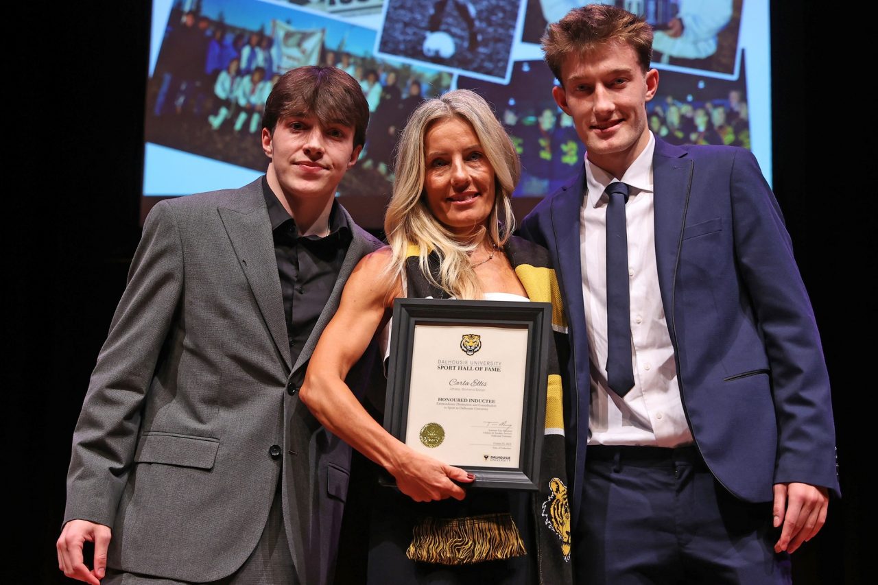 A woman wearing a black and gold scarf holds a framed certificate standing between two young men on stage at an event.