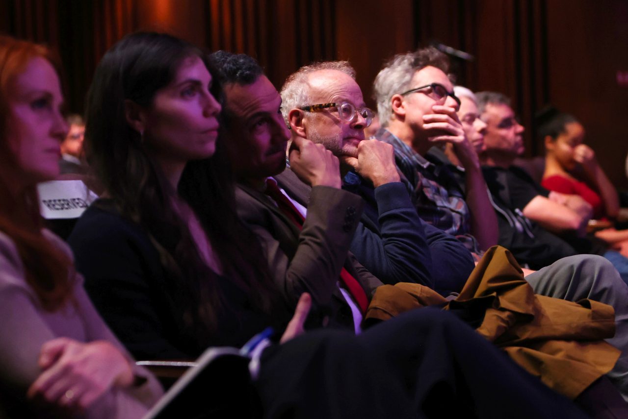 A group of people sit in a darkened room, facing forward, with some resting their heads on their hands and looking attentively at something out of frame. The lighting is dim, with a warm glow illuminating the faces of some individuals in the foreground and mid-ground.