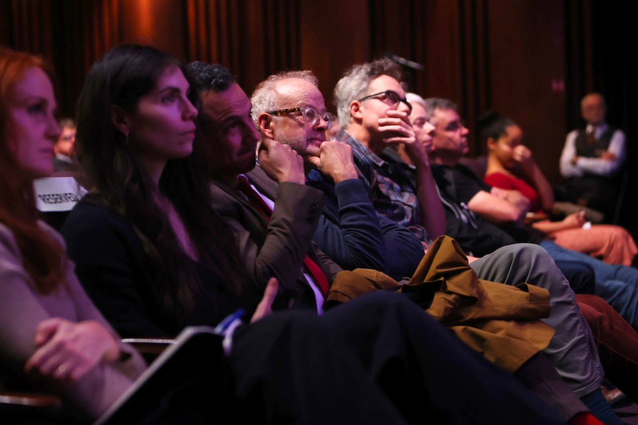 A group of people sit in a darkened room, facing forward, with some resting their heads on their hands and looking attentively at something out of frame. The lighting is dim, with a warm glow illuminating the faces of some individuals in the foreground and mid-ground.