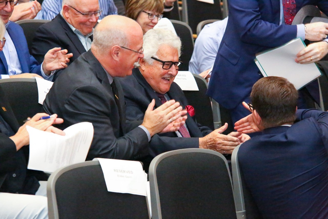 People dressed in business formal attire sitting in auditorium seats clapping and shaking hands.