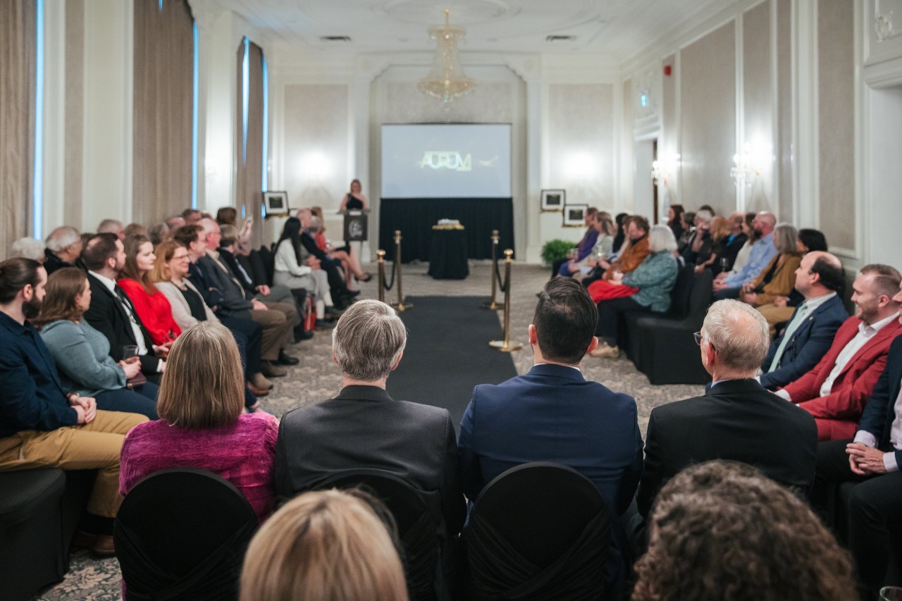 People seated in a horseshoe-shape around a narrow black carpet and video screen at the front while a woman stands at a podium at a semi-formal event.