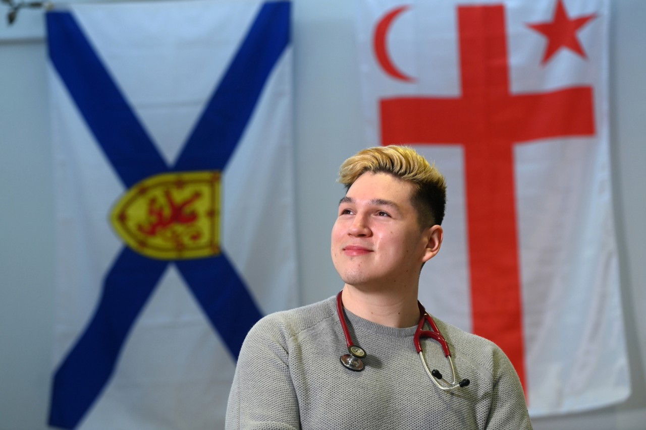 Sylliboy looking in the distance with a stethoscope draped around their shoulders and the Nova Scotia and Mi'kmaq Grand Council flags in the background.