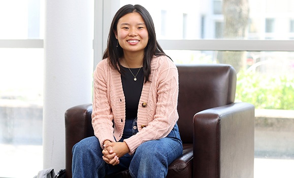 Michelle Wang is sitting in a brown leather chair, wearing a pink cardigan and jeans, smiling at the camera.
