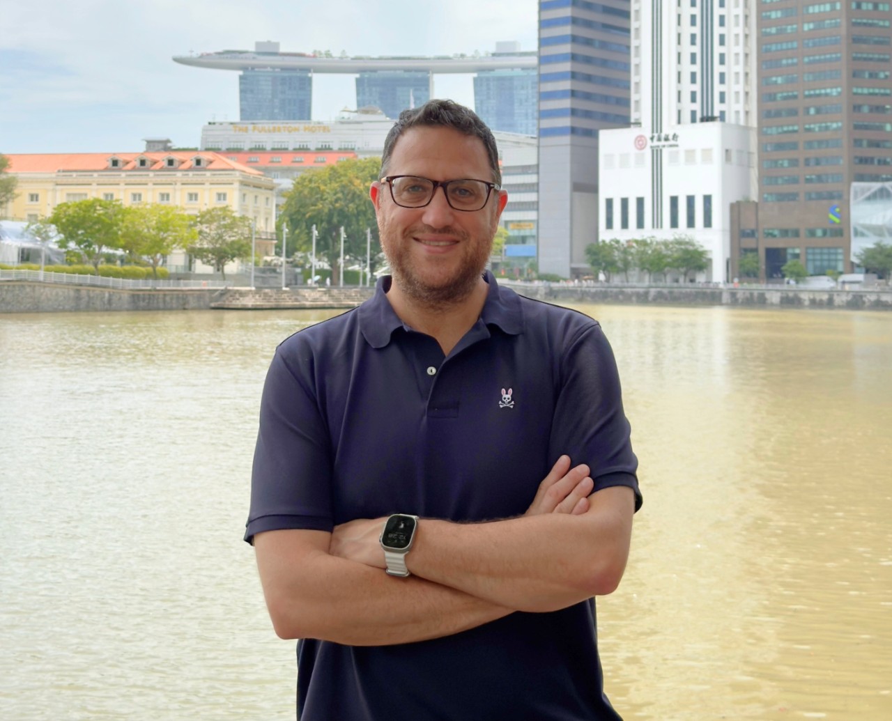 Alon Sobol smiles with his arms crossed while standing in front of a lake in fine weather with skyscrapers behind him.  