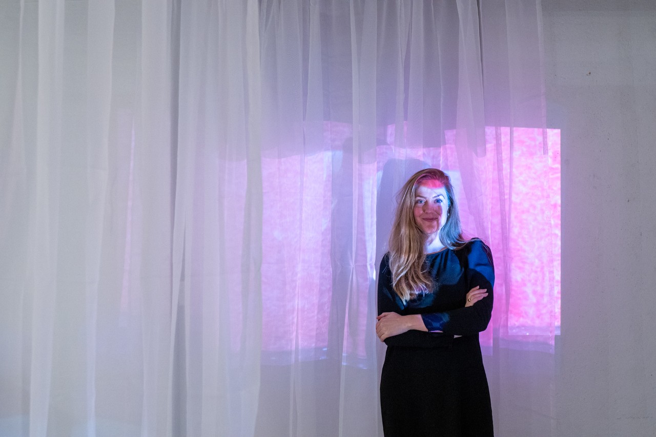 Paulette Cameron standing with her arms crossed casually against a sheer white curtain in a square of purple light.