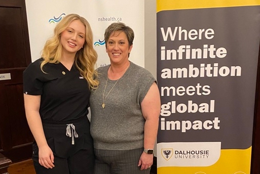 A daughter and mother stand smiling together at an indoor event with banners behind them.