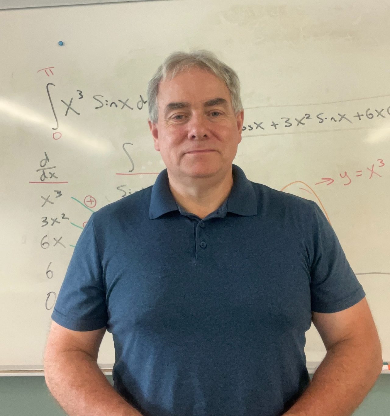Frank Brown standing in a classroom smiling slightly in front of whiteboard.
