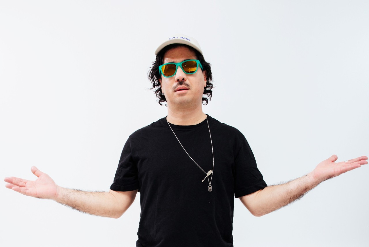 A man in a black t-shirt and light, loose pants with sunglasses and a hat stands with arms outstretched to the sides.