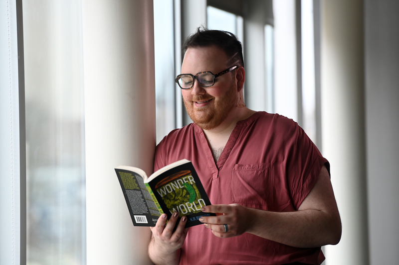 A non-binary person with a beard and black glasses is wearing a red shirt and leaning against a white post. They are smiling and looking at the open book in their hands, which is black and green and titled "Wonder World."