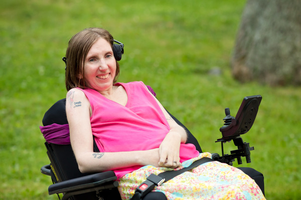 A thin white woman wearing a pink sleeveless shirt sits in a motorized wheelchair, smiling at the camera with her hands clasped in her lap.