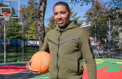 Rodney Small stands on an outdoor basketball court in a green hoodie sweatshirt, holding a basketball and smiling.