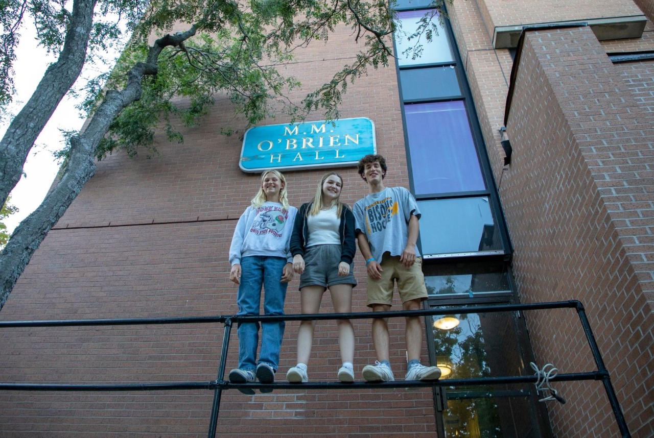 Three students stand on the railing outside a tall brick residence building dressed for warm weather and a leafy tree over top.