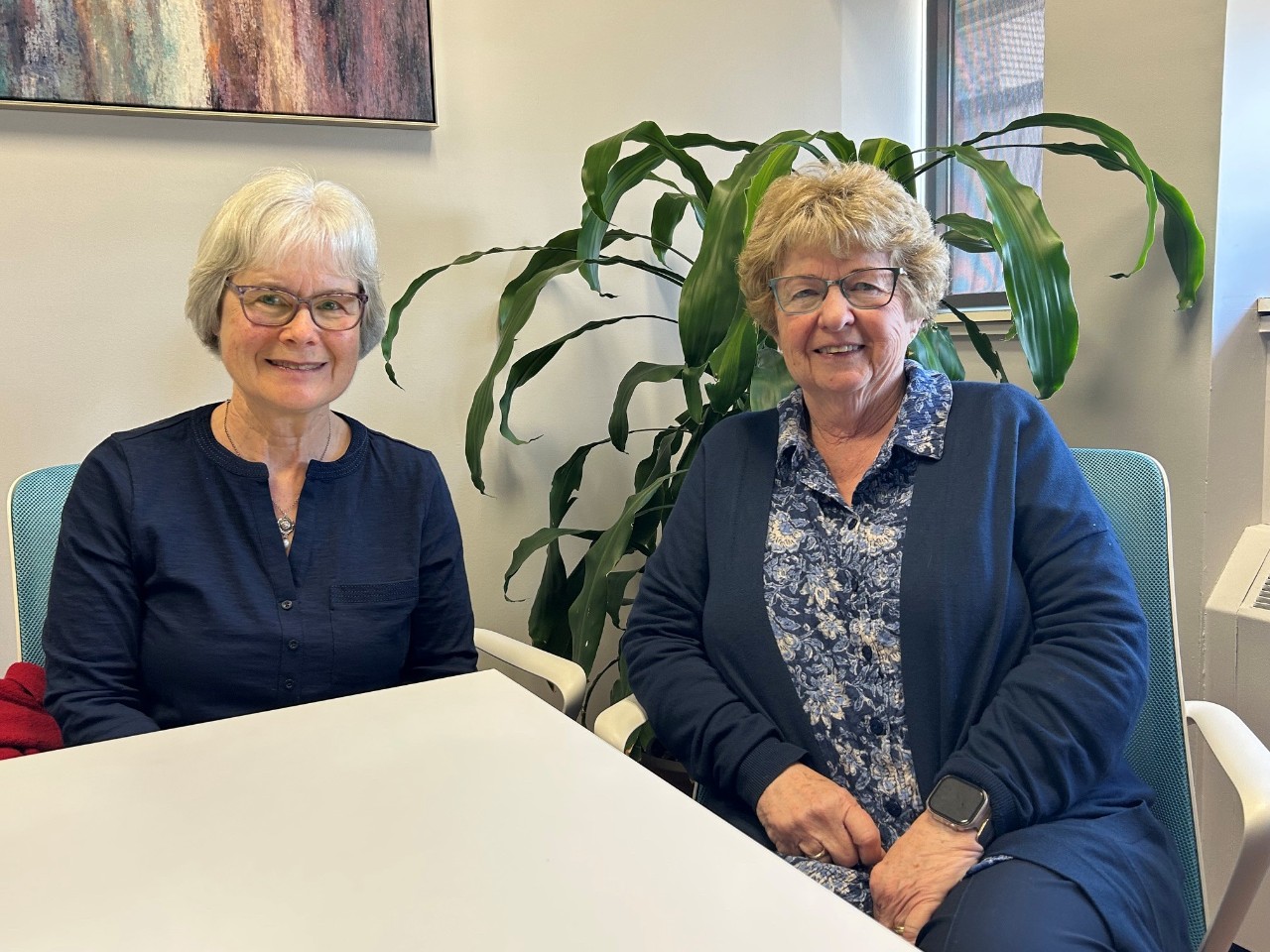 Two women seated at a table smiling in a small bright office space with a large plant.