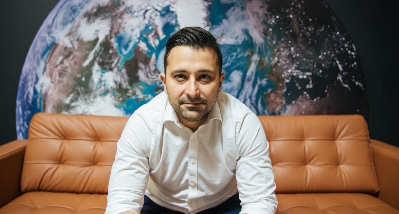 Engineer Arad Gharagozli sits on a couch in front of a wall with a large image of planet Earth.