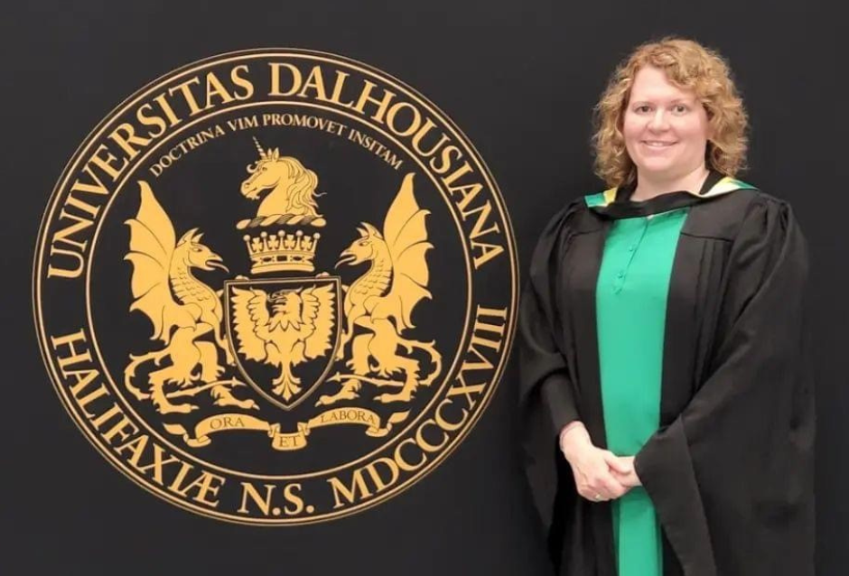 MacQueen stands with hands clasped dressed in a Convocation gown next to a backdrop with the Dalhousie University Seal. 