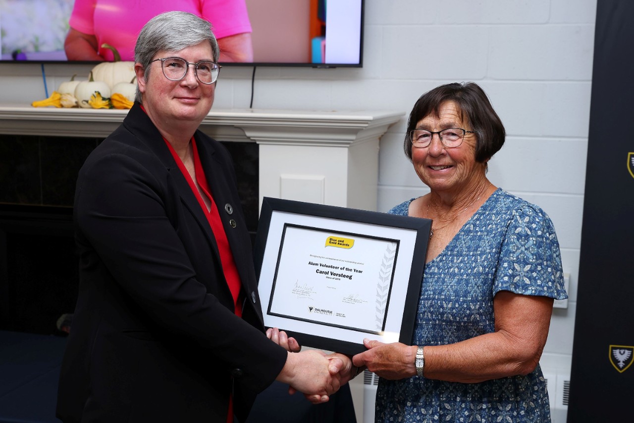 Two women shake hands, smiling at an event with a framed certificate between them.