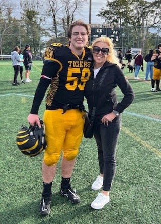 A woman with blonde hair wearing casual black-coloured clothing and sunglasses has her arm around a student in football uniform on the field.