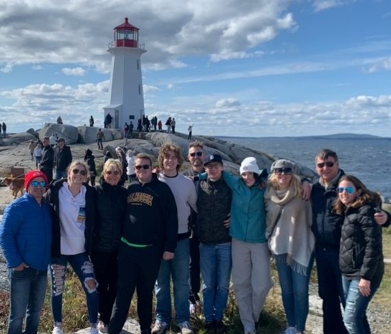 Twelve people stand in a row with arms around each other at Peggy's Cove with the lighthouse in the background.