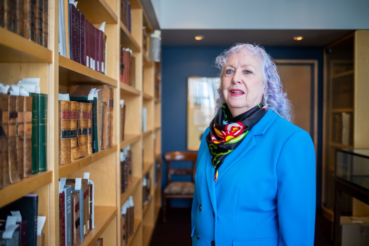 Lynch is wearing a blue jacket and colourful scarf standing facing a shelf of books. 