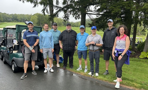 A group of eight people dressed for golf stand in a row alongside a golf course with several parked golf carts.
