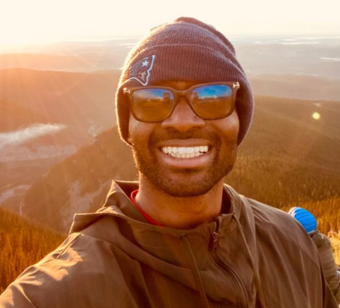 A male-presenting person wearing a toque and sunglasses smiles outdoors with a mountain vista in the background. 
